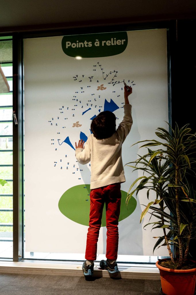 Un enfant relie des points qui formeront un dessin sur un tableau. Il est sur la pointe des pieds, de dos.