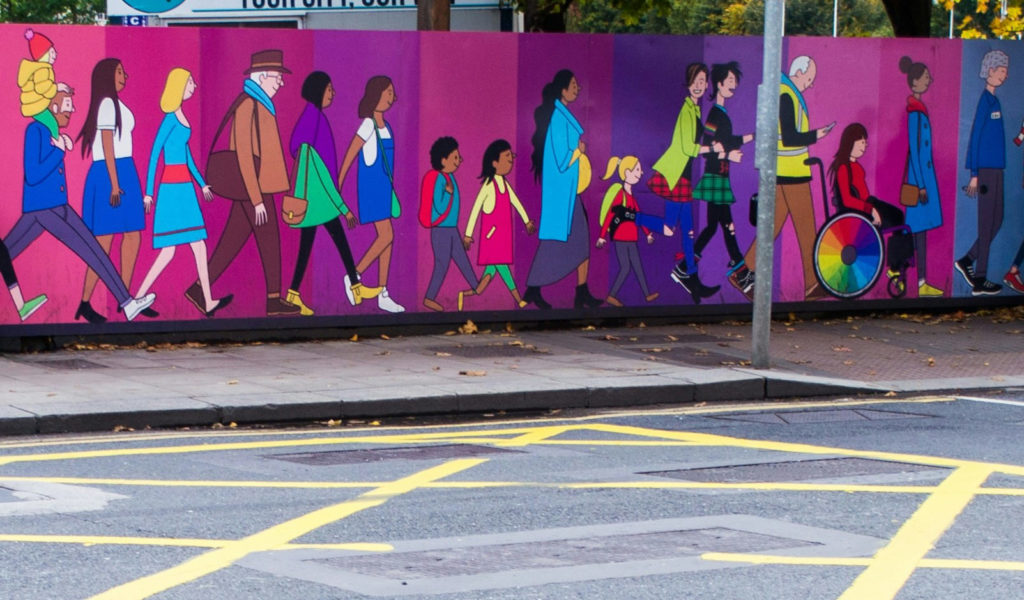 Panneaux peints le long d'un trottoir. L'illustration représente des personnages, dans leur diversité, en train de marcher les uns à la suite des autres.