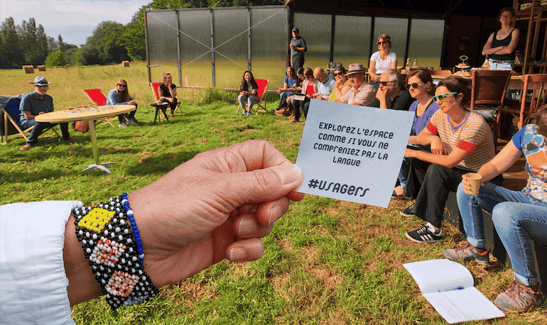 Un groupe est assis dans l'herbe. Au premier plan, une main tient un papier où il est inscrit "explorez l'espace comme si vous ne compreniez pas la langue #usagers"