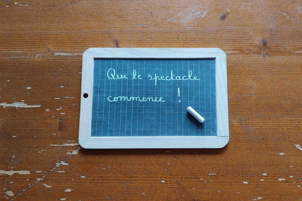 Une ardoise, posée sur une table en bois avec une craie dessus, sur laquelle il est écrit avec une écriture enfantine : Que le spectacle commence !