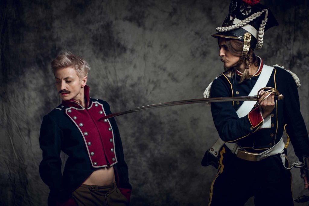 Une femme blonde aux cheveux courts avec une moustache et un costume de cavalier laissant voir son ventre, menacée par une épée tenu par une autre femme habillée également en cavalier.