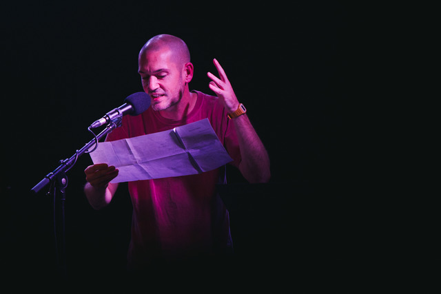 Un homme lit un texte face à un micro. Salle plongée dans le noir, lumière sur lui.