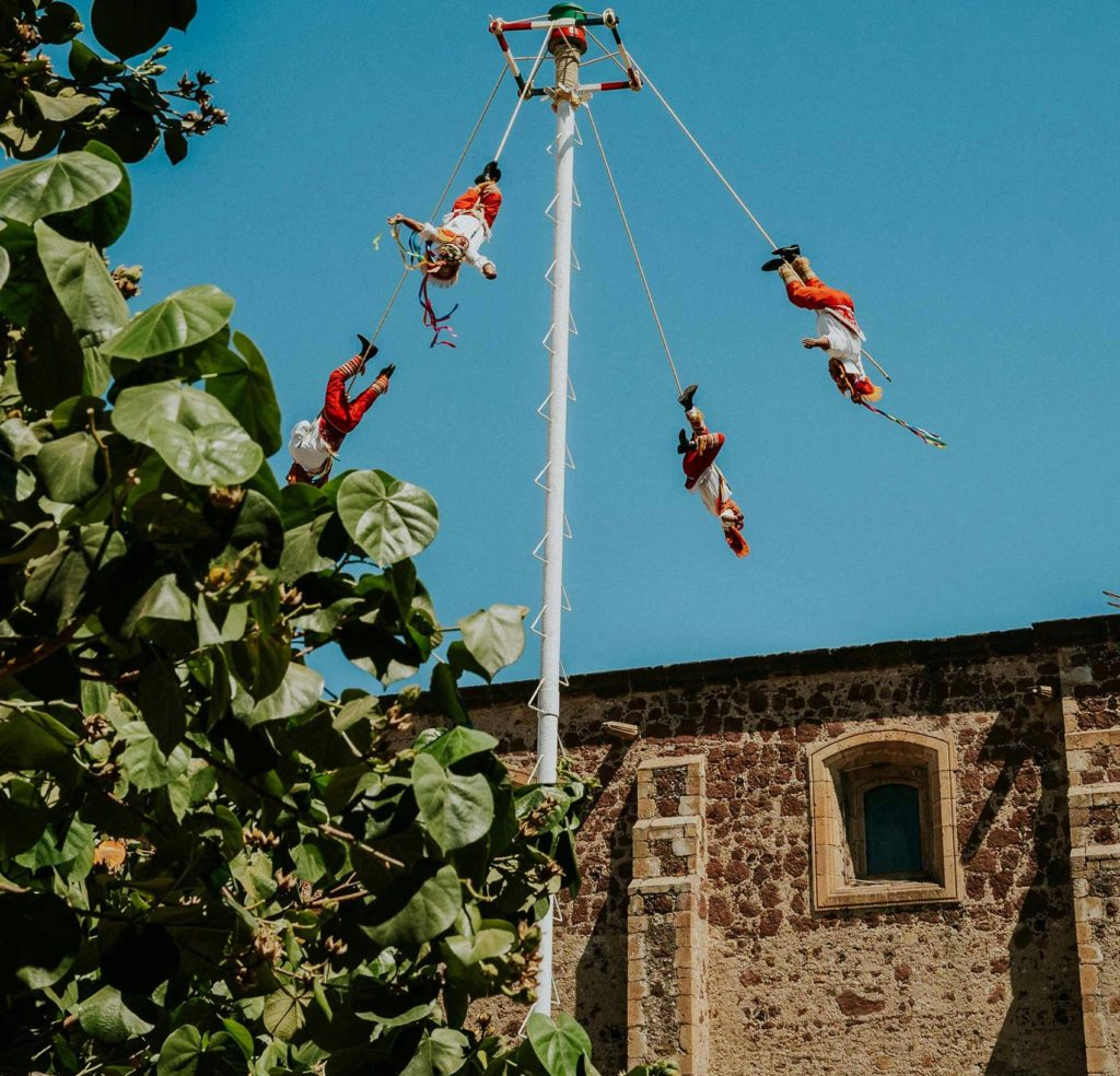 Un mât et des acrobates suspendus. Une église et des feuillages.